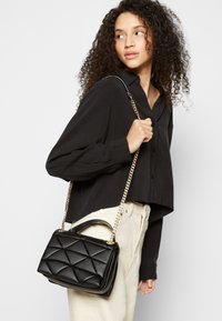 Sac à main noir matelassé avec un design structuré, bandoulière en chaîne dorée et une poignée supérieure, assorti à une chemise boutonnée noire et un pantalon de couleur claire.