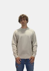 Sudadera beige de manga larga, cuello redondo y puños acanalados. Combinada con jeans azules desgastados. Sutil logo de la marca en el cuello.