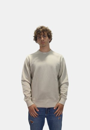 Beige genser med lange ermer, rund hals og ribbede mansjetter. Brukt med slitte blå jeans. Diskret merkevarelogo ved kragen.