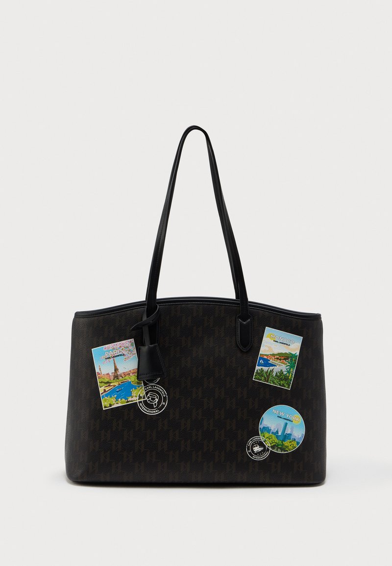 Czarna torba tote w ciemnym odcieniu brązu z subtelnym wzorem i długimi czarnymi uchwytami, ozdobiona naszywkami o tematyce podróżniczej przedstawiającymi Paryż, Monako i Nowy Jork.