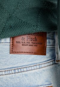 Groene gebreide stof bedekt gedeeltelijk een bruine leren label op lichtblauwe denim. Het label heeft reliëfbranding en stiksels.