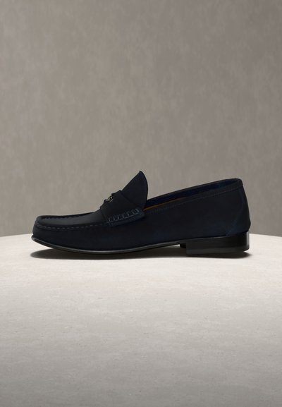 Σκούρο μπλε δερμάτινο παπούτσι τύπου loafer με χαμηλό μαύρο τακούνι, μαύρη σόλα και διακοσμητικό μεταλλικό στοιχείο στο επάνω μέρος, τοποθετημένο σε φωτεινή επιφάνεια.