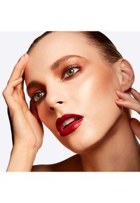 Close-up of a face featuring radiant skin, green eyes, smoky bronze eyeshadow, and bold red lipstick. Smooth texture and natural lighting highlight the features.