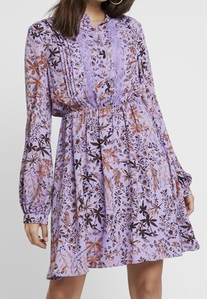 Kvinde iført en lilla blomsterkjole med lange ærmer, blonder detaljer foran og elastik i taljen, stående mod en ensfarvet baggrund.