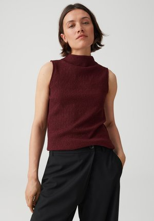 Donna con capelli corti e castani che indossa un top smanicato bordeaux texturizzato e pantaloni neri, con una mano in tasca su sfondo semplice.