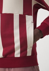 Main dans la poche d'un sweat-shirt rayé bordeaux et blanc cassé avec poignets et ourlet côtelés visibles sur un bras à la peau foncée.