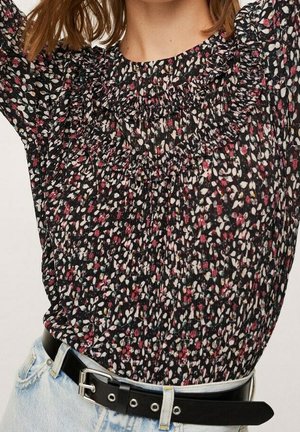 Femme portant un chemisier smocké noir à motifs floraux, rentré dans un jean bleu clair avec une ceinture noire ornée d'œillets argentés et d'une boucle.