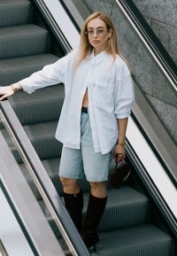 Chemise blanche oversize à boutons, shorts en denim bleu clair, bottes marron jusqu'aux genoux, tenant un petit sac rond bordeaux. Textures lisses.