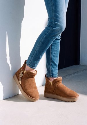 Une personne portant un jean bleu et des bottines en daim beige se tient contre un mur blanc, un pied incliné et posé sur le talon.