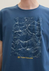 Plava pamučna majica s grafičkim printom vodnih uzoraka u nijansama plave i bijele, s oznakom "TOM TAILOR" u žutoj boji na dnu.