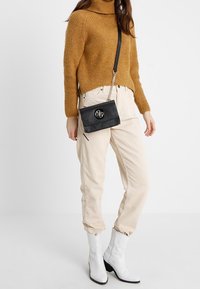 Sort læder crossbody taske med kædetræk og et rundt logo. Båret med en sennepsfarvet turtleneck sweater og beige corde pants. Hvide støvler.