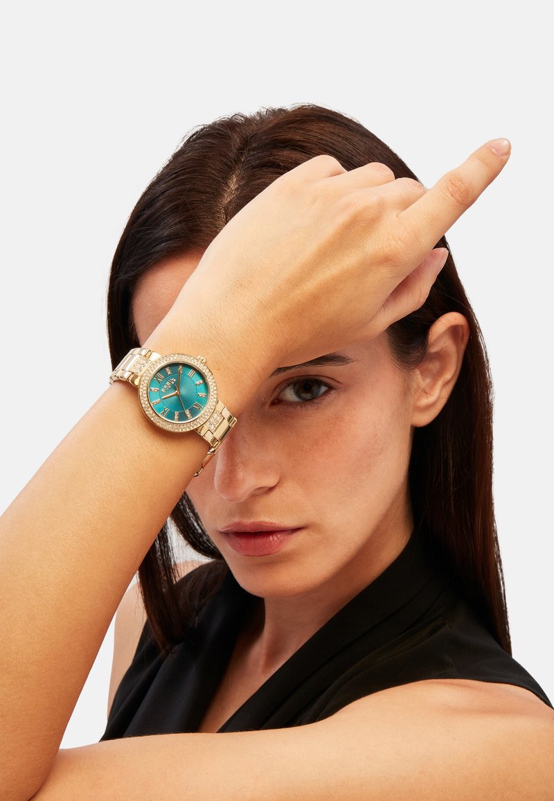 Montre dorée avec un cadran turquoise, des index en chiffres romains, et une lunette ornée de cristaux, accompagnée d'un bracelet en maillons métalliques.