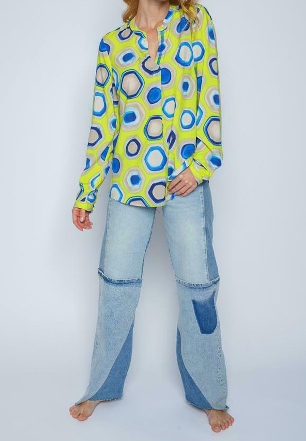 Femme portant un chemisier vert vif à motifs d’hexagones bleus et blancs et un jean large en patchwork, debout pieds nus sur un fond uni.