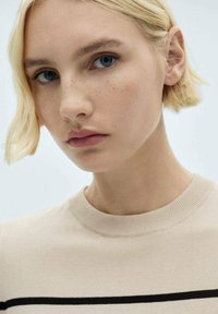 Beige knit sweater with a crew neckline, featuring black horizontal stripes. Model has short, wavy blonde hair and stands against a light background.