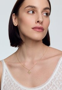 Złoty naszyjnik z zawieszką w kształcie wielkiej litery "M", delikatny łańcuszek, noszony na białym, teksturowanym topie na ramiączkach. Miękkie oświetlenie podkreśla detale.