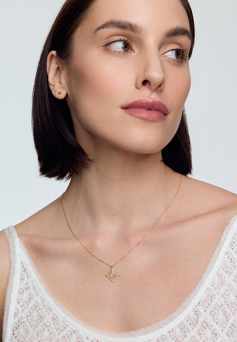 Złoty naszyjnik z zawieszką w kształcie wielkiej litery "M", delikatny łańcuszek, noszony na białym, teksturowanym topie na ramiączkach. Miękkie oświetlenie podkreśla detale.