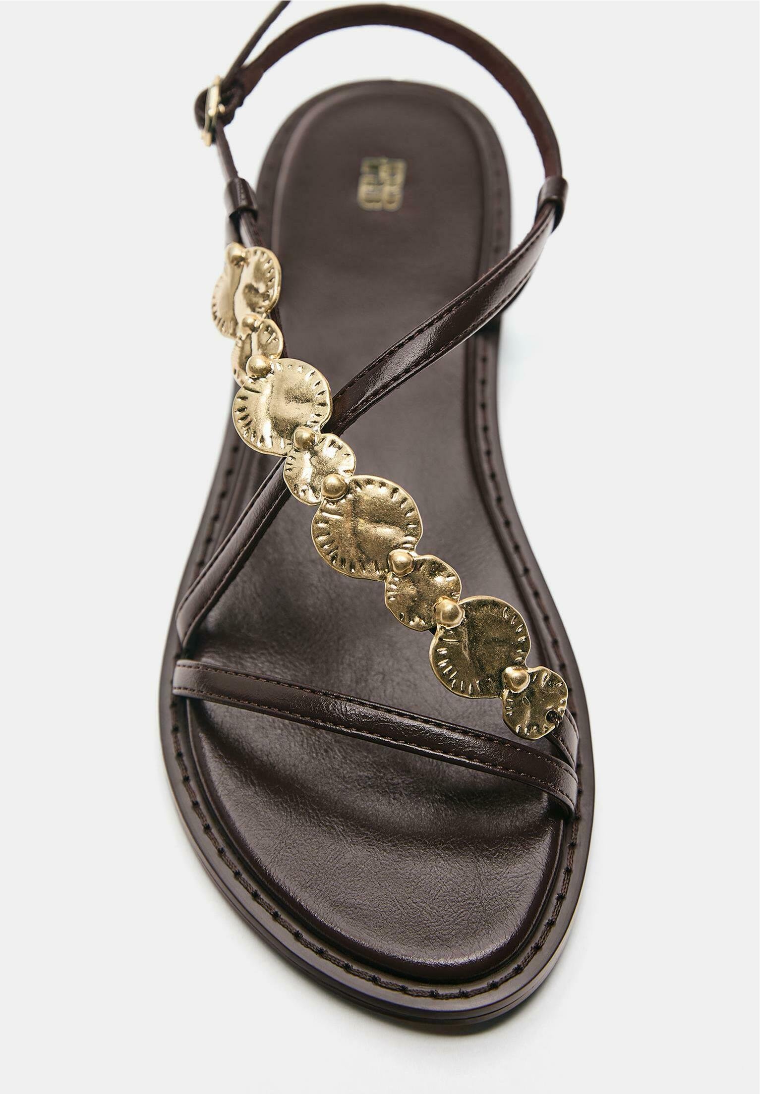 Temno rjava usnjena sandal z ozkimi trakovi in zlato dekorativnimi okroglimi kovinskimi okrasnimi elementi vzdolž zgornjega traku.