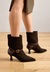 Brown suede ankle boots with a folded cuff, pointed toe, and a thin heel. Features a brown leather strap with a gold buckle accent.