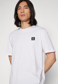Tricou alb din bumbac, cu guler rotund, mâneci scurte și un mic patch cu logo-ul Timberland de culoare neagră pe zona din stânga a pieptului.