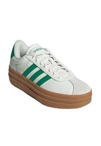 Vit sneaker med gröna ränder och logotyp, som har ett slätt läderöverdrag och en sula av gummigummi. Snörningen synlig framtill.