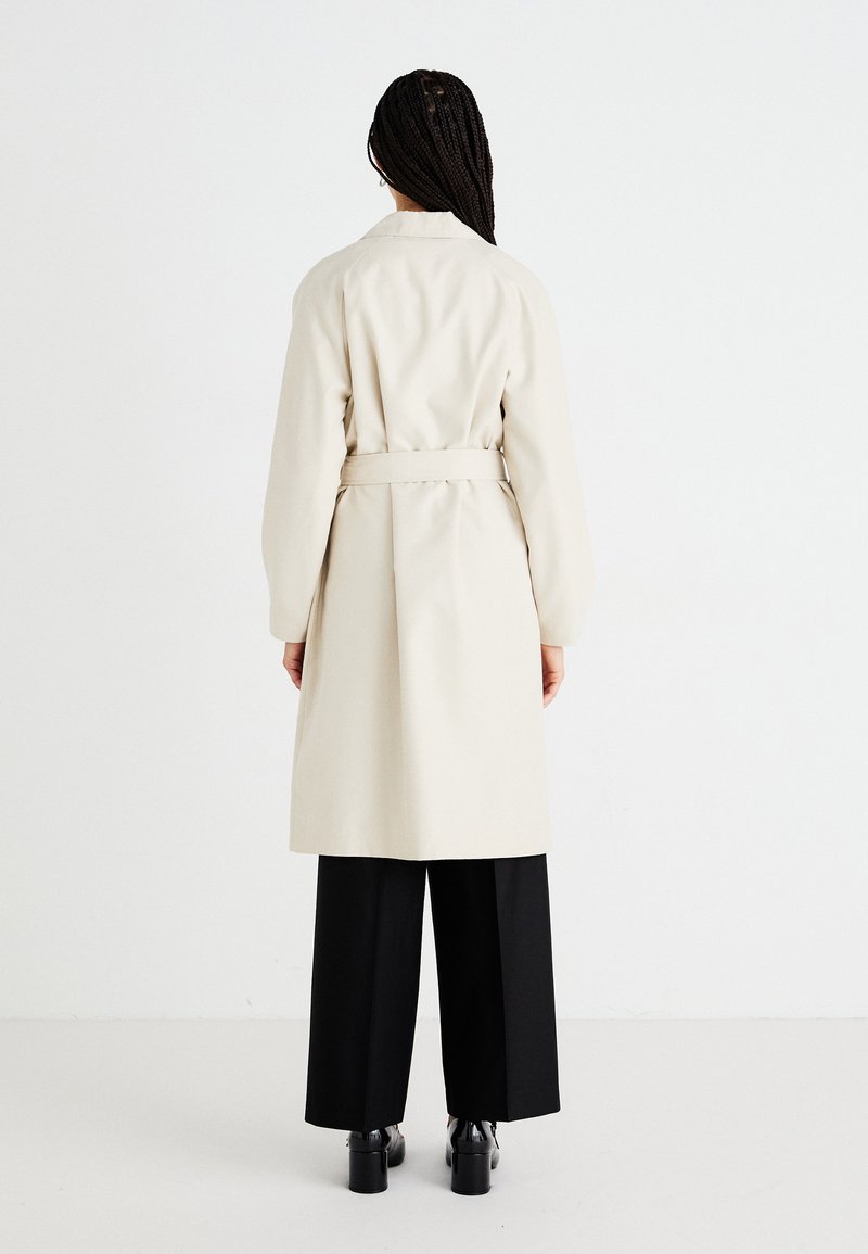 Beżowy długi płaszcz z paskiem w talii, posiadający długie rękawy i prosty krój. Nosi się go na czarne szerokie spodnie i buty na obcasach.
