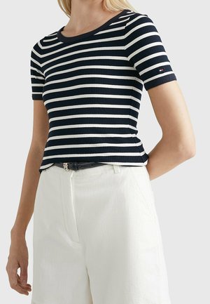 Femme portant un haut en maille à manches courtes rayé bleu marine et blanc avec un short blanc taille haute et une ceinture noire.