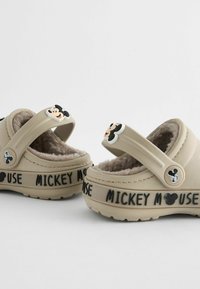 Beige kengät pehmeällä vuorilla, joissa on Mickey Mouse -koristeet ja "MICKEY MOUSE" -teksti sivussa. Säädettävä hihna tukevan istuvuuden takaamiseksi.