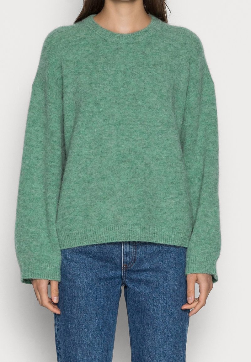 Femme portant un pull doux vert pelucheux à manches longues et un jean denim bleu, debout devant un fond clair uni.