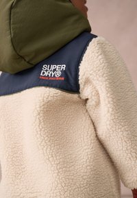 Sherpa flísová bunda s krémovou textúrou, námorníckou hornou časťou, zelenou kapucňou a červeným vyšitým logom. Má voľný strih a neformálny dizajn.
