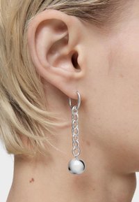 Pendiente de aro de plata con un diseño de eslabones de cadena, que presenta un colgante redondo y liso. El pendiente cuelga por debajo del lóbulo de la oreja en un modelo.