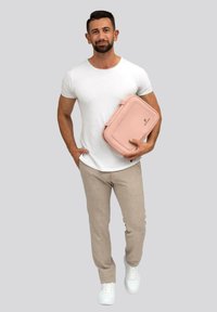Lichtroze neplederen tas vastgehouden door een persoon in een wit shirt en beige broek. De tas heeft een gladde textuur en een minimalistisch ontwerp.