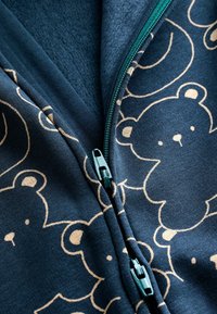 Navyblaue Stoffjacke mit beigen Bärenmustern und Reißverschluss. Der Reißverschluss ist türkisfarben mit einem glatten Lauf, das strukturierte Fleecefutter ist sichtbar.
