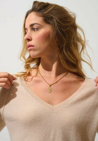 Goldenes Anhänger-Halskette an einer dünnen Kette, getragen über einem hellbeigen V-Ausschnitt-Pullover mit geripptem Kragen und weichem Material.