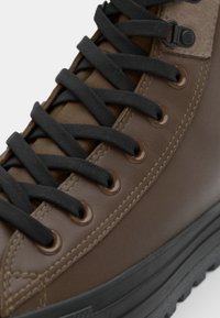 Smeđi kožni visoki sneakers s crnim vezicama, koji ima metalne rupice, teksturirani jezik i debeli crni gumeni potplat.
