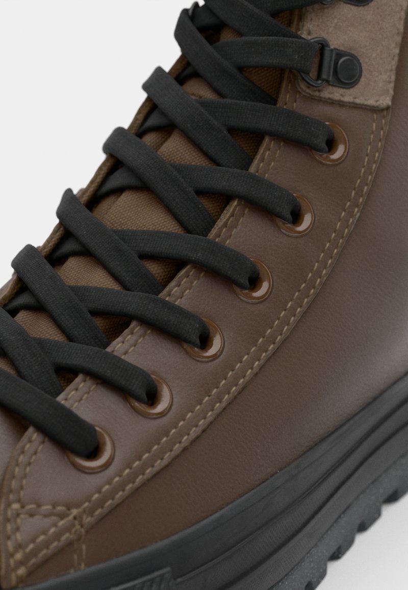 Sneaker alta in pelle marrone con lacci neri, dotata di occhielli metallici, una linguetta testurizzata e una spessa suola in gomma nera.