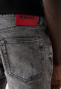 Jeans de denim gris con una textura desgastada, con una etiqueta de cuero roja que dice "HUGO" en la parte trasera de la cinturilla y bolsillos traseros estándar.