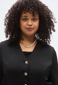 Fekete blézer arany virágos gombokkal, fekete felsőre viselve. Gyöngy és aranylánc nyaklánc. Göndör haj keretezi az arcot.