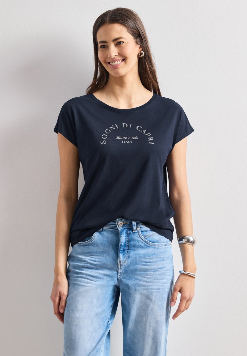 Smiling woman wearing navy blue t-shirt with "Sogni di Capri" text and light blue jeans standing against plain white wall.