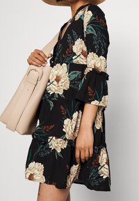 Abito nero floreale con maniche a tre quarti, orlo arricciato e borsa beige. Presenta grandi fiori color crema e foglie verdi.