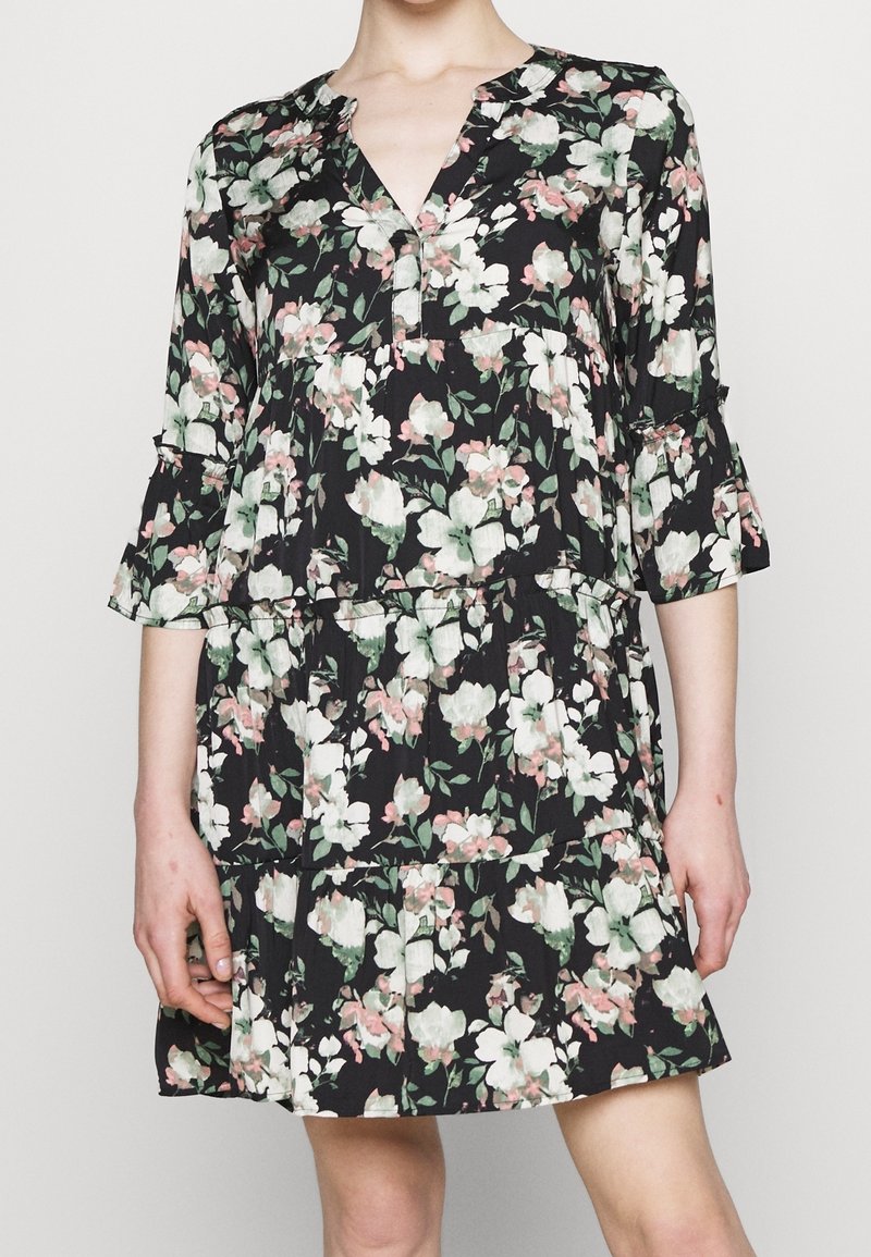Personne portant une robe noire à hauteur du genou à imprimé fleuri blanc et rose, manches trois-quarts et encolure en V, debout devant un fond uni.