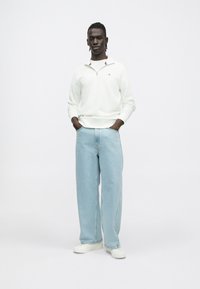 Fehér cipzáras pulóver csíkos gallérral, világoskék, bő szárú farmerrel és fehér cipővel; a modell zsebbe dugott kézzel pózol.