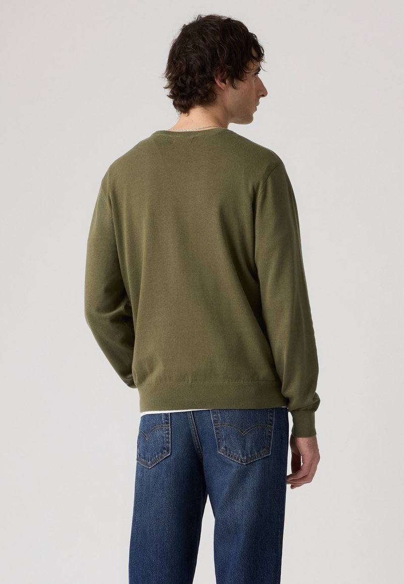 Man with short curly hair wearing an olive green sweater and blue jeans, standing and facing away against a plain background.