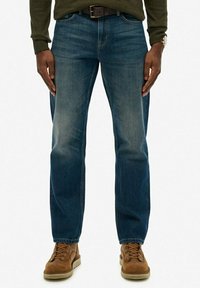 Męskie niebieskie dżinsy z denimu, prosta nogawka, średnie pranie z delikatnym wyblaknięciem. W zestawieniu z brązowymi butami na sznurówki, dopełnione ciemnym paskiem.