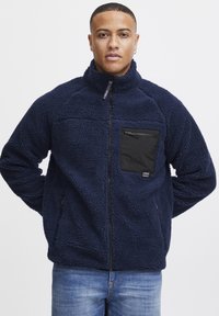 Navy blue fleece jacket with a textured surface, full zip, high collar, and a black zippered chest pocket. Features side pockets.