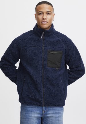 Chaqueta de forro polar azul marino con una superficie texturizada, cierre completo, cuello alto y un bolsillo en el pecho con cremallera negra. Incluye bolsillos laterales.