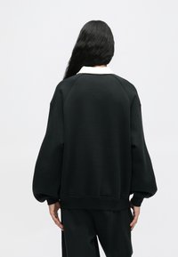 Άτομο με μακριά σκούρα μαλλιά, φορώντας μια χαλαρή μαύρη μπλούζα με λευκό γιακά, στέκεται με την πλάτη γυρισμένη σε απλή απαλή φόντο.