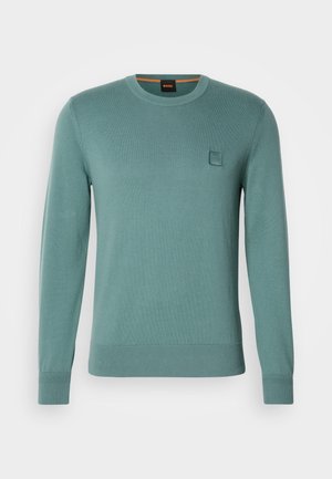 Lyse teal strikket genser med rund hals, lange ermer, ribbestrikkede mansjetter og midje. Har en liten firkantet logo-lapp på brystet.