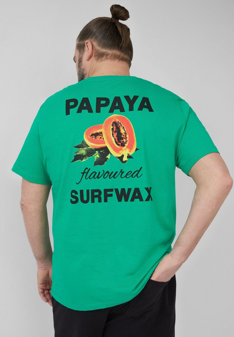 Groene katoenen t-shirt met een afbeelding van gesneden papaja en zwarte tekst: "PAPAYA-smaak SURFWAX". Korte mouwen en een ontspannen pasvorm.