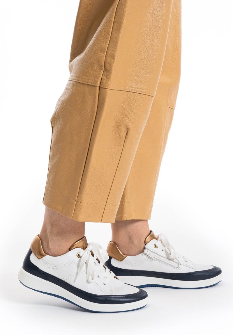 Weiße und marineblaue Sneakers mit braunen Akzenten, die über ein elegantes Design, eine runde Zehenpartie und eine Schnürung verfügen. Getragen mit hellbraunen, cropped Lederhosen.