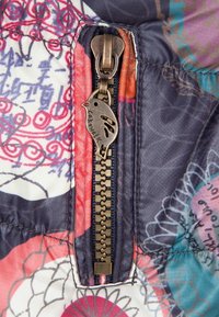 Kleurvolle, gemusterde stof met een opvallende ritssluiting en een decoratieve vogelsleutel. De rits heeft een messingkleur en een rechthoekig ontwerp van tanden.